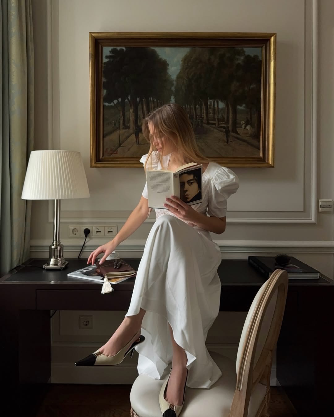 Mujer de vestido blanco leyendo sentada sobre un escritorio, bajo un cuadro